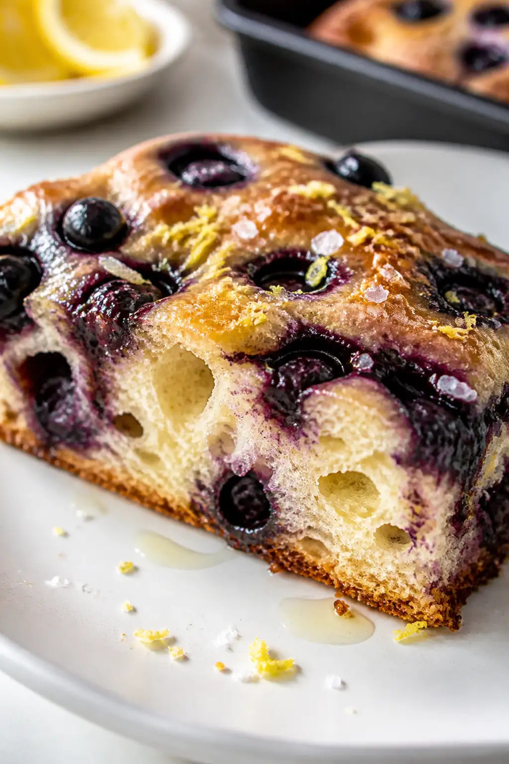 Close-up detail of a warm slice of lemon blueberry focaccia on a simple white plate, showing the air